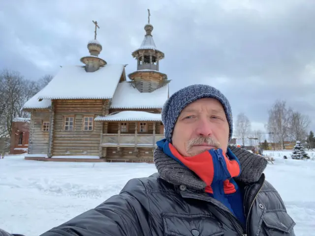 Усатый мужчина в зимней вязаной шапке на фоне зимнего пейзажа делает селфи