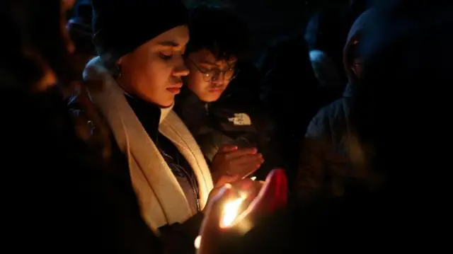 夜間の屋外に人々が集まっている。それぞれが防寒具を身に着け、両手にろうそくを持っている。祈るように見つめている顔が、ろうそくの光で照らされている