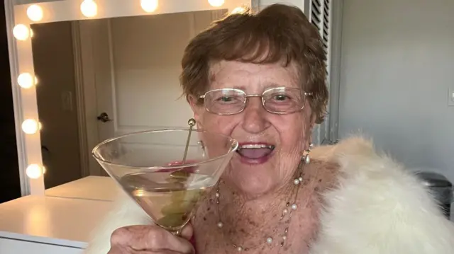 Lillian Droniak, de 96 anos, segura uma taça de martini com azeitonas. Ela sorri enquanto usa óculos, uma estola de pele e um colar com caimento fluido; seu cabelo curto e avermelhado está arrumado, e há um espelho com moldura iluminada ao fundo.