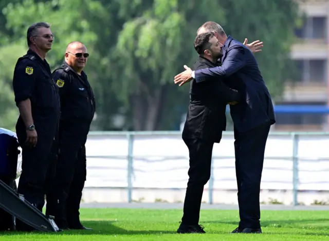 Зеленский и Мерц обнимаются, на них смотрят двое мужчин в форме.