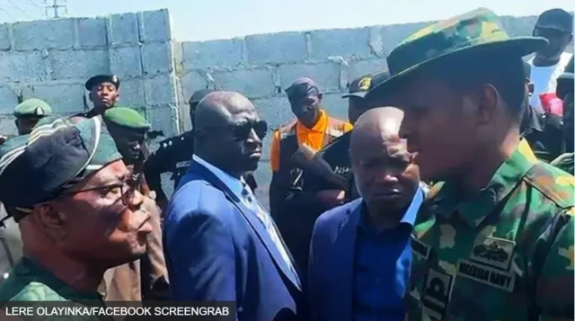 Wike ń woju ọmọ ológun to wa ninu aṣọ ológun tí tíì ṣe aláwọ̀ èwe. Ọ̀pọ̀ èèyàn yí àwọn méjèèjì ká.