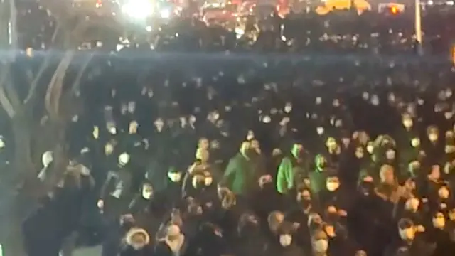 Một nhóm đông người tụ tập trên đường phố vào ban đêm. Hầu hết đều đeo khẩu trang và nhiều người trùm mũ áo khoác. Những luồng sáng chiếu tới làm rực lên một bóng cây, phía xa là những ánh đèn sáng rực.