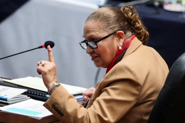 La fiscal general de Guatemala, Consuelo Porras, sentada en una mesa con documentos, alza el índice de su mano izquierda. 