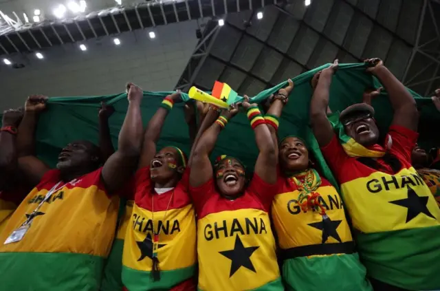 Üzerlerinde Gana renkleri (yeşil, sarı ve kırmızı) ve ortasında siyah bir yıldız bulunan tişörtler giyen, kadın ve erkek beş futbol taraftarı. Heyecanlı görünüyorlar ve ellerinde bayrağın bir parçasını başlarının üzerinde tutuyorlar. Bir stadyumdalar.
