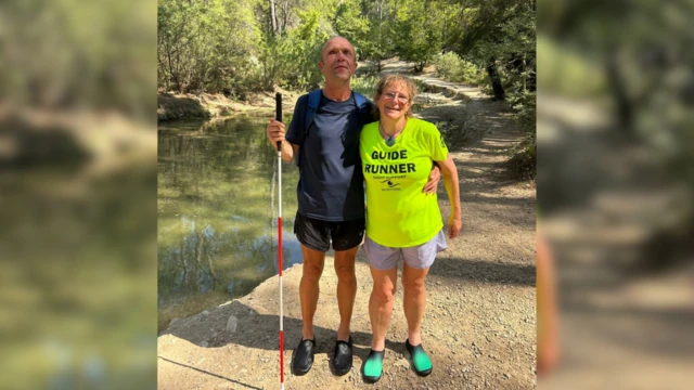Keith y Katy posan en la acera junto a un río en el campo. Keith sostiene un bastón de movilidad extendido en la mano derecha. Katy lleva una camiseta luminosa con la inscripción "GUIDE RUNNER". Árboles de hoja caduca bordean la orilla del río, creando sombras en la acera al fondo. Ambos sonríen mientras el sol ilumina el rostro de Katy.