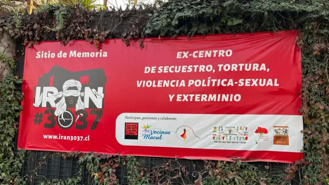 Gros plan d'une bannière rouge à l'extérieur des murs du site commémoratif, anciennement connu sous le nom de Venda Sexy. La bannière porte une inscription blanche en gras sur laquelle on peut lire : « ancien centre de détention, de torture, de violence politico-sexuelle et d'extermination ». La bannière comporte également un dessin de la maison en noir, le nom Iran #3037 et le dessin d'une femme aux yeux bandés. 