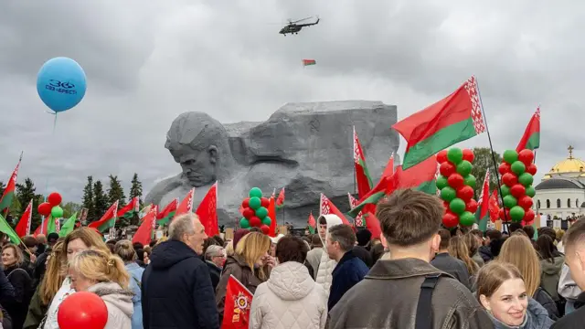 Велике зібрання людей перед монументом із величезною кам’яною скульптурою. У натовпі багато червоно‑зелених прапорів і кульок тих самих кольорів, над головами видно гелікоптер, який несе прапор. Небо затягнуте хмарами, поруч стоїть будівля із золотим куполом.
