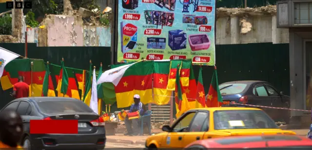 Des véhicules passent sur une route avec à l'arrière-plan un homme portant une casquette, assis avec autour de lui des drapeaux camerounais.