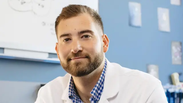 Ben Rein’in baş fotoğrafı. Açık kahverengi kısa saçları ve kahverengi sakalı var. Kapalı bir mekânda, beyaz laboratuvar önlüğü ve mavi-beyaz kareli bir gömlekle görüntülenmiş.
