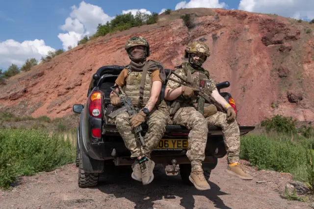 Hai nữ binh sĩ Ukraine thuộc Lữ đoàn Đổ bộ Đường không độc lập số 25 “Sicheslav”, mang biệt danh “Malibu”, 34 tuổi (trái), và “Little One”, 21 tuổi (phải), tạo dáng chụp ảnh trên một chiếc xe bán tải, ngày 31/5/2024 tại tỉnh Donetsk, Ukraine. “Malibu” và “Little One” là các trắc thủ hệ thống máy bay không người lái của đại đội trinh sát thuộc Lữ đoàn Đổ bộ Đường không độc lập số 25 “Sicheslav”. Nhiệm vụ của họ là phát hiện hầm trú ẩn của đối phương, khí tài, nhân lực và phối hợp dẫn bắn vào các mục tiêu này.