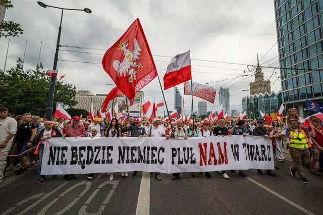 Вулична хода в центрі Варшаві. Попереду колони учасники тримають довгий білий банер з чорним і червоним написом польською: "Не буде німець плювати нам в лице". Над натовпом майорять численні польські прапори, а також великі червоні стяги з білим орлом.