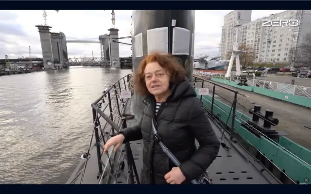 70-річна жінка з вʼюнким каштановим волоссям стоїть на палубі пришвартованого підводного човна. Позаду видно воду, підйомний міст у піднятому положенні та міські будівлі вздовж набережної.