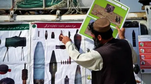 تصویر یک کارمند اجتماعی در حال تشریح انواع مهمات منفجر ناشده به مردم. او به عکس نارنجکی که در تابلویی در روبه‌رویش است اشاره می‌کند. 