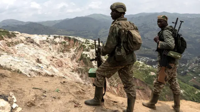 Deux hommes en tenue de combat et armés se tiennent au sommet d'une colline gardant un site minier dans l'est de la République démocratique du Congo, avec des collines ondulantes au loin.