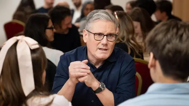 黒いフレームのめがねに、紺のシャツを着たスターマー氏が、若い男女と向かい合って椅子に座り、話をしている。両肘をテーブルに乗せ、両手を組んでいる