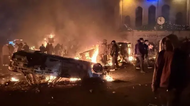 Группа людей в ночное время на площади в Тегеране на фоне опрокинутых автомобилей и костров