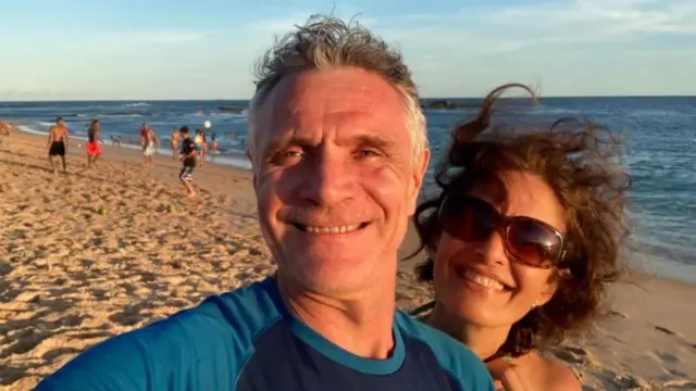 Fotografia colorida mostra casal de homem e mulher brancos sorrindo com uma praia e o mar ao fundo