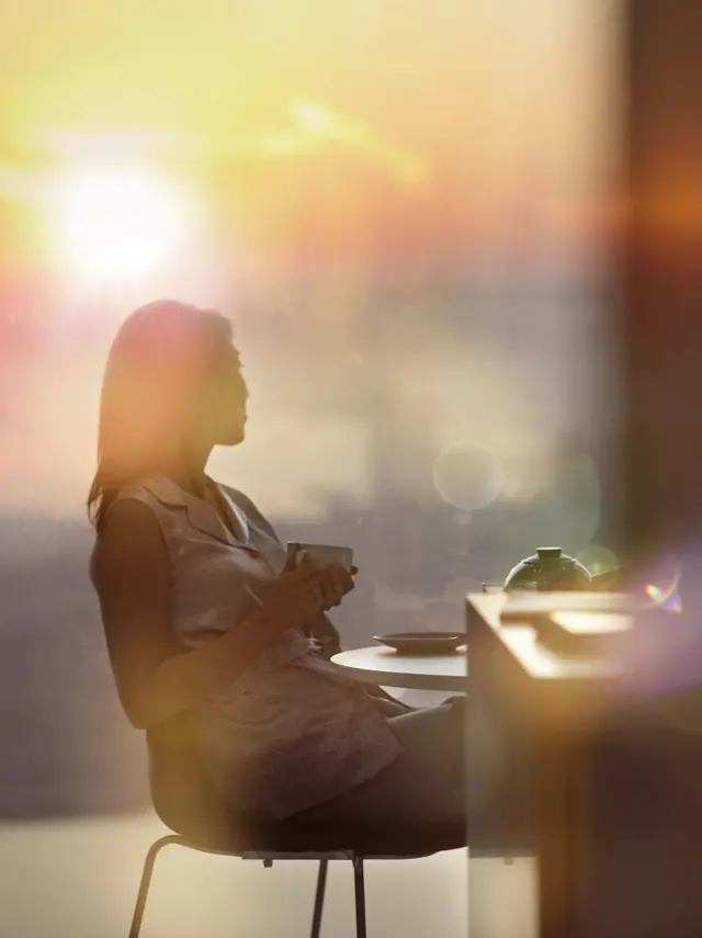 Жінка з чашкою кави сидить за столом і дивиться вбік, фото в мʼяких тонах і сонячних бліках 