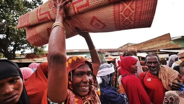 A woman wey lift mat up as she dey smile to di camera, a woman wear red hijab beside anoda man wey wear red polo