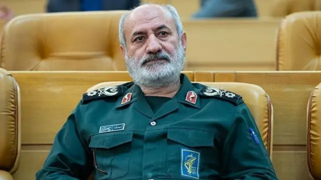 محمد کاظمی در یک سالن سخنرانی با لباس نظامی روی صندلی نشسته است