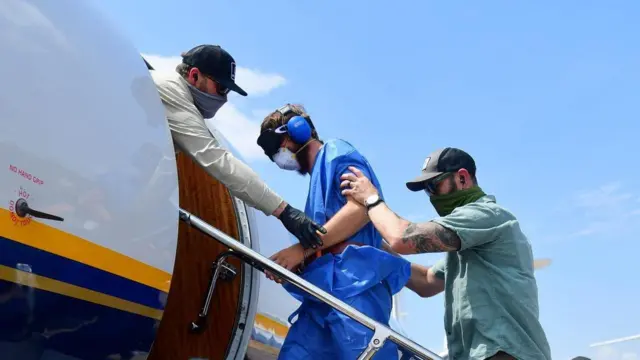 Umwe muri izi mfungwa yurizwa indege imusubiza iwabo. Ari hagati y'abagabo babiri, yambaye imyenda y'ubururu. 