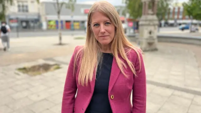 Uma mulher de cabelos longos e loiros, vestindo blazer rosa, está em uma praça vazia, olhando para a câmera.