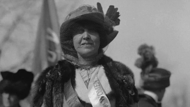 Katharine McCormick wearing a sash showing the word 'Women' with a banner being held in the background