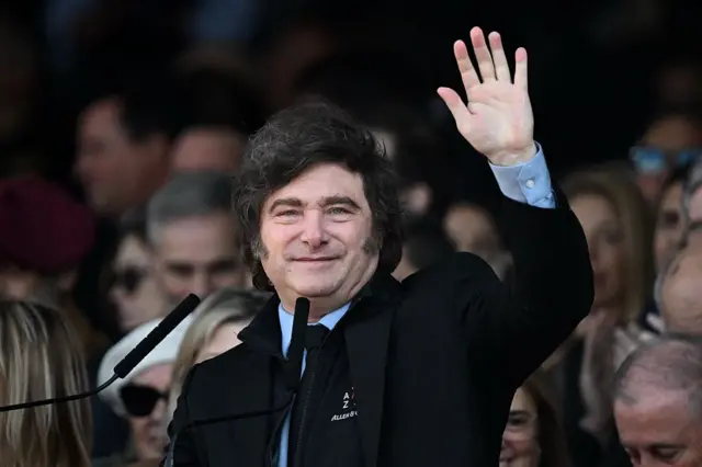 Javier Milei saludando con un traje oscuro y camisa color celeste.