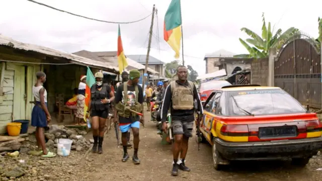 One man wear shorts and a bullet-proof vest. e dey wit two odas wey wear di same dress.