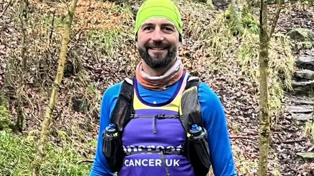Un hombre sonríe a la cámara. Lleva un sombrero amarillo brillante, una bufanda naranja, una camiseta azul de manga larga y un chaleco morado. Está de pie en un bosque.