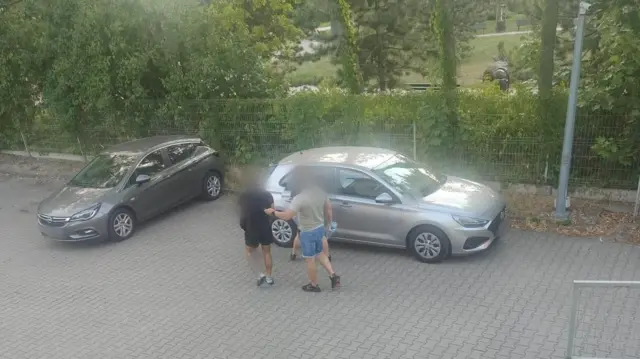 Mężczyzna ubrany w beżowy t-shirt i niebieskie spodenki prowadzi innego mężczyznę, ubranego w czarny t-shirt i czarne spodenki, w kierunku srebrnego samochodu. Ich twarze są zamazane.
