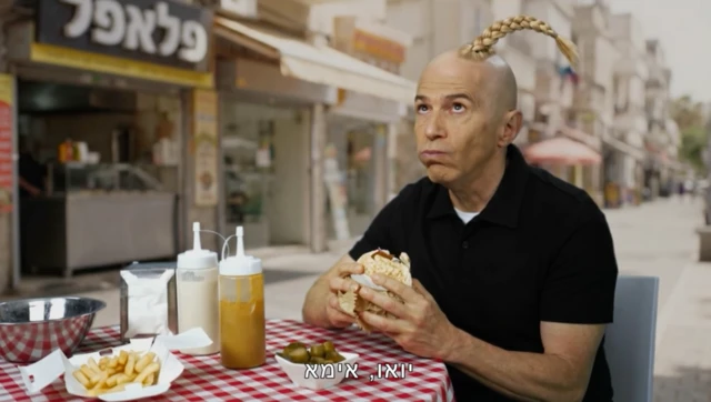 Bir adam restoran masasına oturmuş yemek yerken saçı sarı tek örgülü komik bir hal almış. 