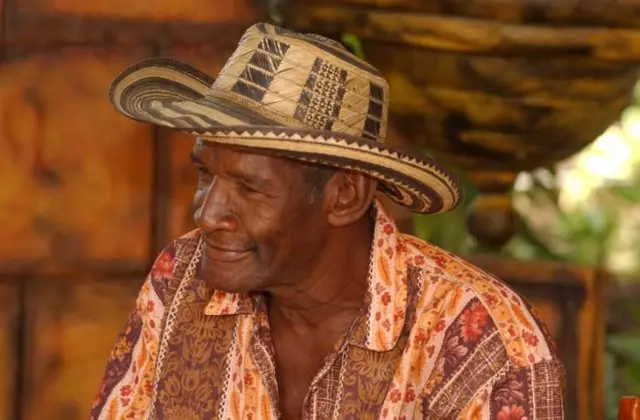 Evaristo Márquez sonriente, con un sombrero típico de tejido de caña y una camisa de colores marrón y naranja. 