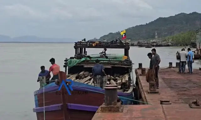 တောင်တန်းနောက်ခံမြင်ကွင်းမှာ ဆိပ်ကမ်းဆိုက်ကပ်ထားတဲ့ သစ်သားသင်္ေဘာ အနီရောင်၊ ခရမ်း အနားကွပ် ပေါ်မှာတင်ဆောင်ထားတဲ့ သစ်တွေနဲ့ ပတ်ဝန်းကျင်မှရပ်နေတဲ့ အမျိုးသားတွေ 