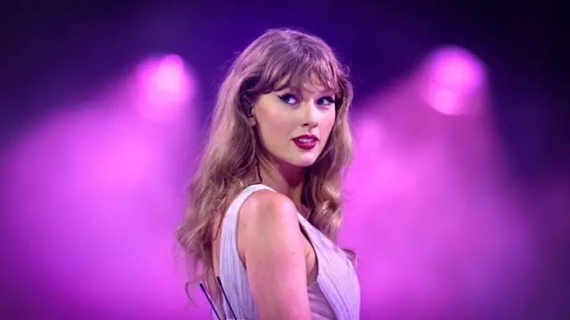 Taylor Swift em close durante uma apresentação, olhando por cima do ombro em direção ao público. Vestindo um figurino lilás e iluminada por refletores em tons de rosa e roxo, ela aparece com expressão atenta, tendo ao fundo o telão do palco, rosa e desfocado, que destaca sua presença em cena.