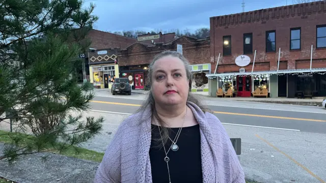 Mulher com cabelo grisalho e casaco lilás, de pé em frente a uma série de lojas, na praça principal de Gainesboro.
