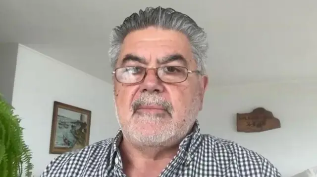 A mid-shot of Jorge Odón as he looks directly into the camera. He has short grey hair and is wearing glasses and a black and white checked shirt. 
