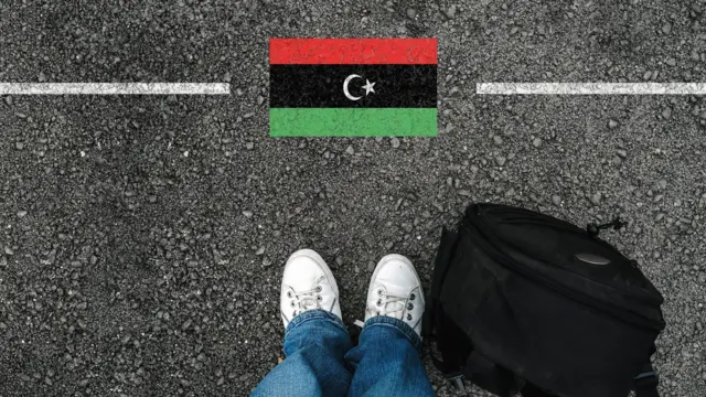 رجل يرتدي حذاءً وحقيبة ظهر يقف على الأسفلت بجوار علم ليبيا والحدود.
