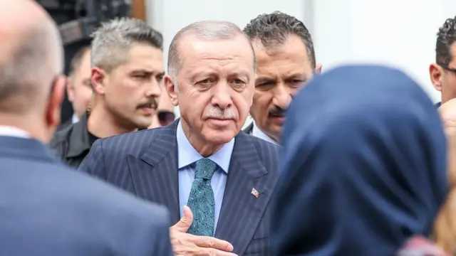 Cumhurbaşkanı Erdoğan, bir seçim mitingi öncesinde taraftarlarını selamlıyor. Arkasında korumalar var, kime baktığı anlaşılmıyor, elini göğsüne götürerek insanları selamlıyor.