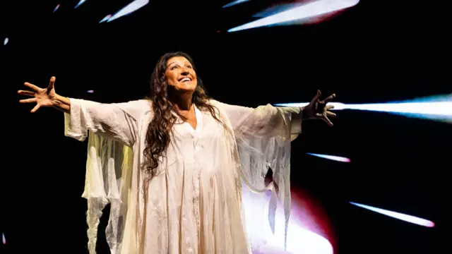 Regina Casé veste uma longa túnica branca de mangas amplas e está no palco com os braços abertos e um sorriso voltado para o alto. Ela usa um microfone de cabeça, enquanto, ao fundo, há projeções de feixes de luz sobre um cenário escuro.