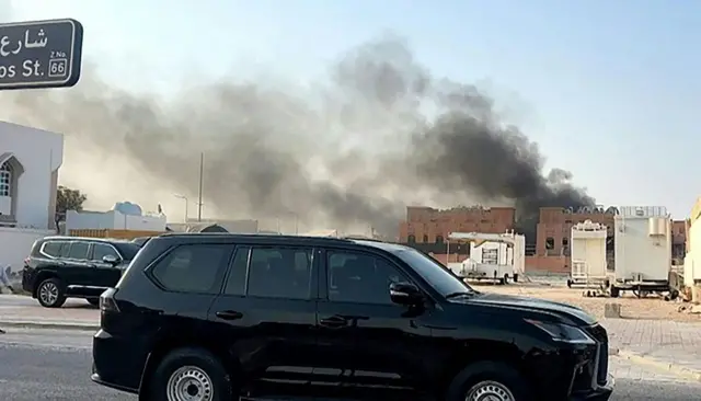 Una camioneta negra transita por una calle de Doha, mientras que al fondo se ven varios edificios y una nube de humo negro saliendo de ellos