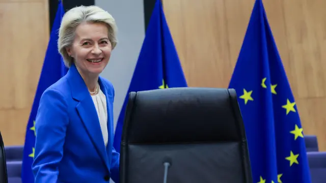 A presidente da Comissão Europeia, Ursula Von der Leyen, uma mulher brancamollybetcabelo curto loiro claro ficando grisalho, com um blazer  azul,mollybetfrente a três bandeiras da união europeia