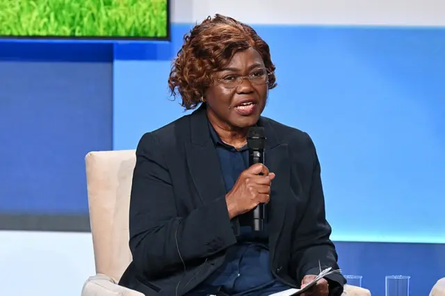 Nialé Kaba porte une veste noire sur une chemise bleue foncée, elle parle dans un micro qu'elle tient dans sa main droite.