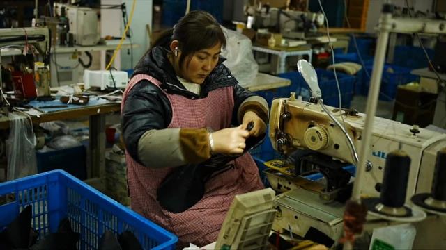 عاملة في مصنع صيني، ترتدي مريولاً أحمر منقط بالأبيض فوق ملابسها، وأمامها ماكينة خياطة وبجانبها سلة زرقاء