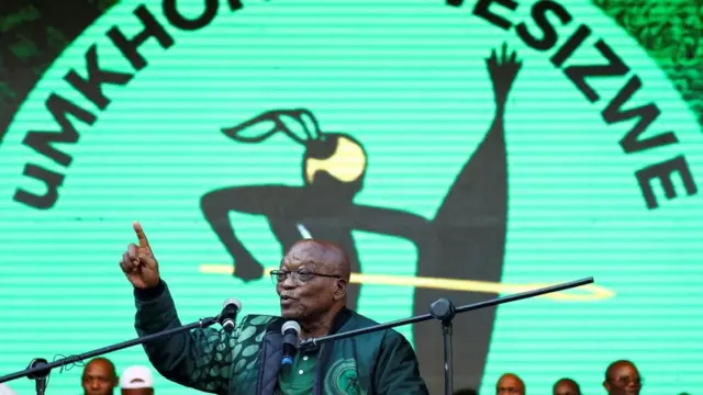 Jacob Zuma s'adresse à la foule lors d'un rassemblement, avec en arrière-plan le logo de son parti : une lance et un bouclier.