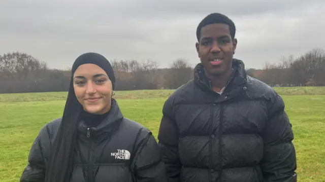 Zainab dey wear a black hijab and a black North Face coat. Jaiden dey wear a black puffer coat. Dem stay side-by-side for one field wit trees in di distance. 