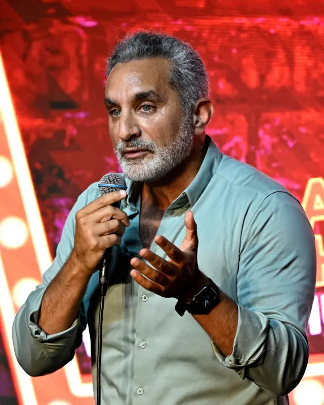 باسم يوسف أثناء تقديمه أحد عروض الستاند أب كوميدي في الولايات المتحدة الأمريكية مرتدياً قميصاً أخضر اللون وساعة سوداء اللون وأمامه مايكروفون.