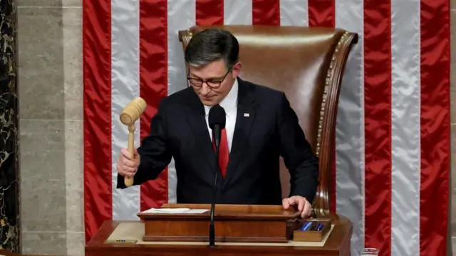 ジョンソン議長は議長席に向かって立ち、法案可決を宣言し、議長用の小づちを右手に持ち振り上げ、まさにこれからたたこうとしている。後ろに星条旗の紅白の線が見える