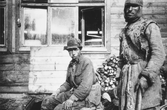 A black and white image on Alderney showing two prisoners of war wearing quilted cotton wool jackets and hats. The one in the centre is sitting, the one on the right standing and holding what looks like a farming tool. Behind them is a wooden hut with open windows.