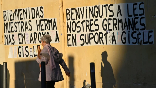 Seorang perempuan berjalan melewati slogan yang bertuliskan "Selamat datang para saudari kami, [yang] datang untuk mendukung Gisele" di tembok kota. 
